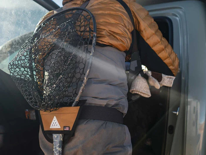 Fisherman wearing a Yakoda Net Holster holding a fishing net with a black rubber mesh and tan leather holder, dressed in outdoor fishing gear near a vehicle.