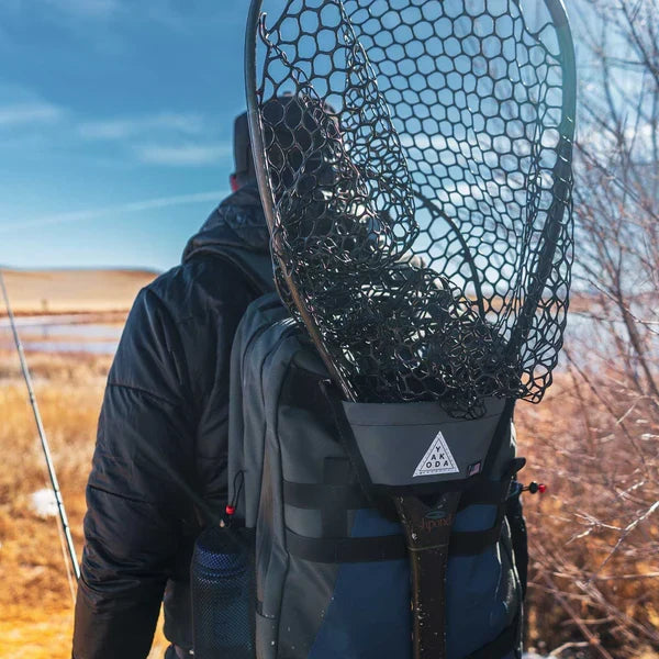 Person wearing outdoor gear carrying a Yakoda Net Holster attached to a backpack, holding a fishing net in a natural lakeside setting, suitable for fishing tackle and gear.