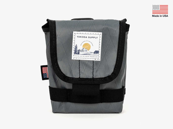 Yakoda Utility Pouch in gray with black trim, featuring a front flap secured by a buckle, a stitched label with outdoor design, and a small American flag tag on the side.