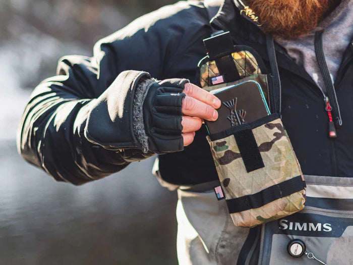 A person wearing outdoor fishing gear places a small item into the camouflage Yakoda Utility Pouch attached to their chest, highlighting its compact and accessible design for fishing tackle use.