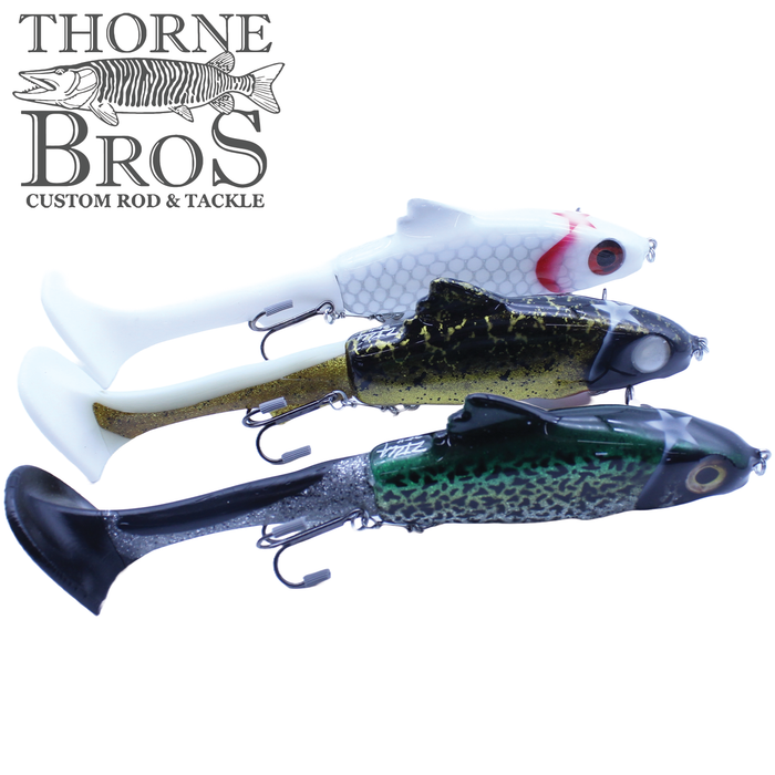 Three detailed soft plastic fishing lures in white, gold, and green colors, each with treble hooks, displayed against a white background featuring the Thorne Bros logo, representing the Zilla Baits Hooker.